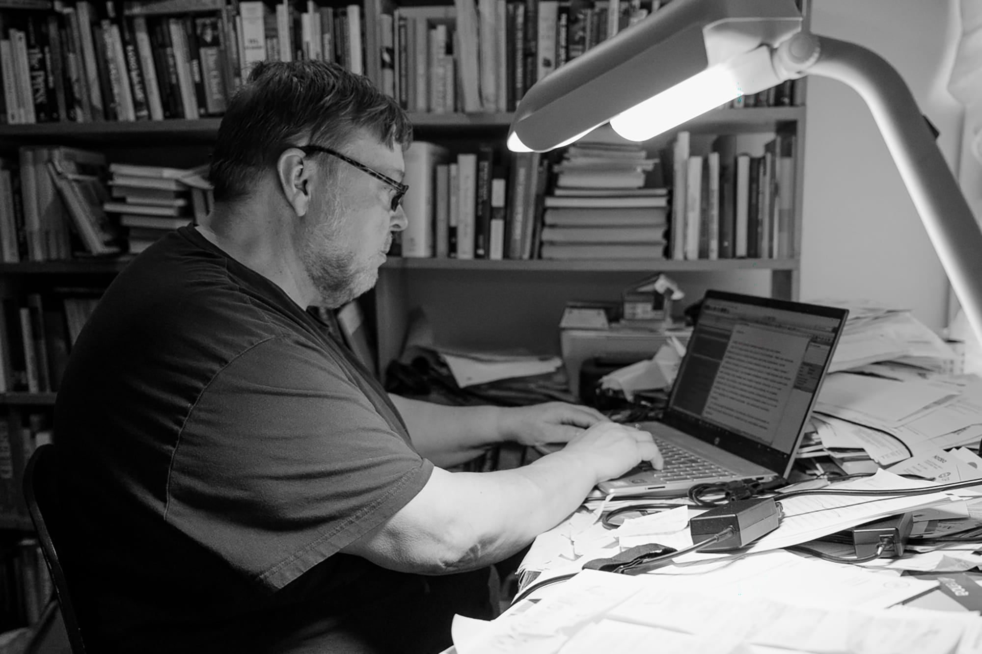 Silmälasipäinen henkilö kirjoittaa työpöydän ääressä.